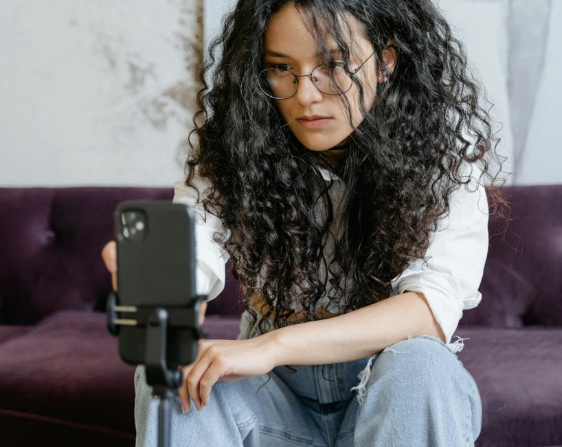 Woman with curly hair vlogging indoors, using smartphone on tripod, seated on couch.